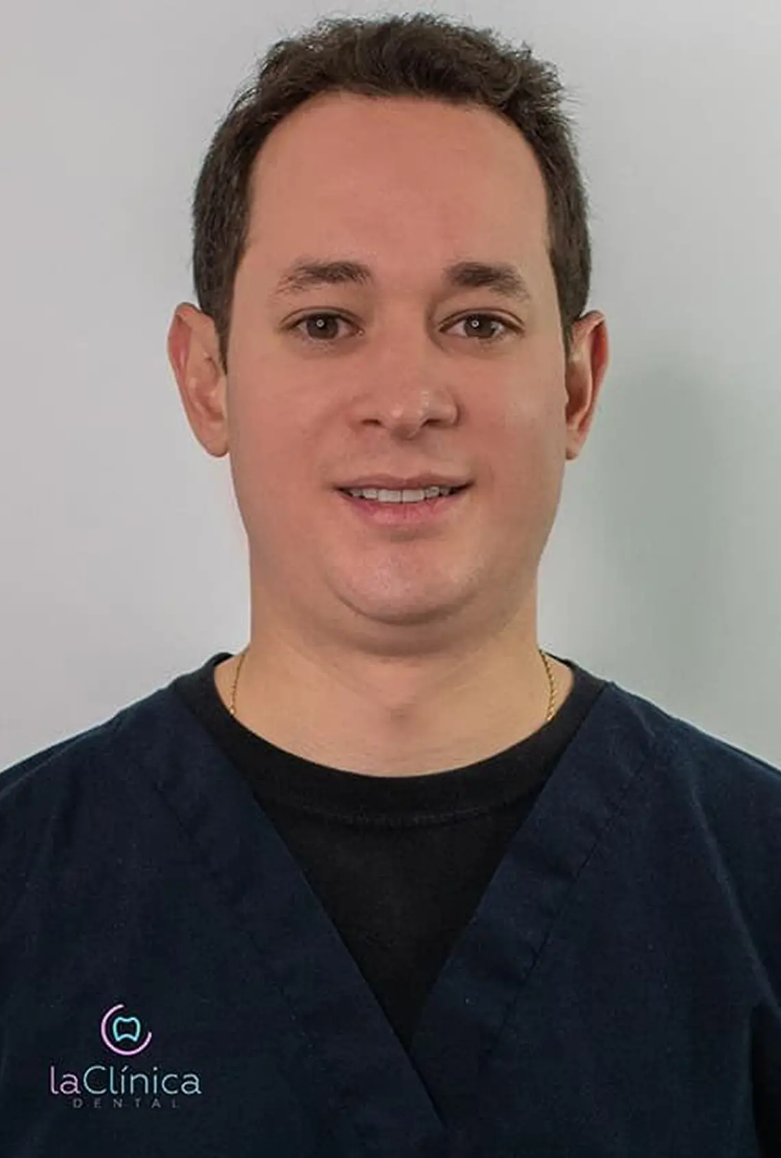 Dentista en La Clínica Dental Roma con uniforme azul, sonriendo frente a fondo blanco, representando atención dental de calidad.