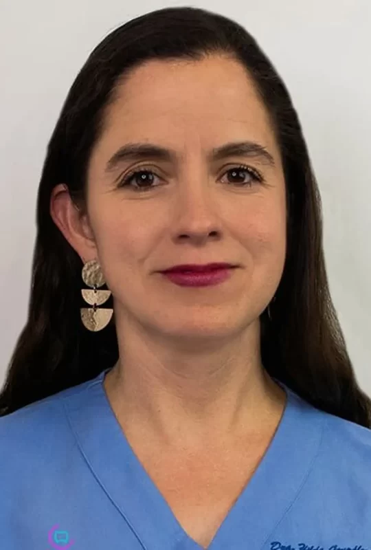 Dr. Hilda G., specialist in Maxillofacial Surgery, wearing a blue dental scrubs and statement earrings, with a neutral background.
