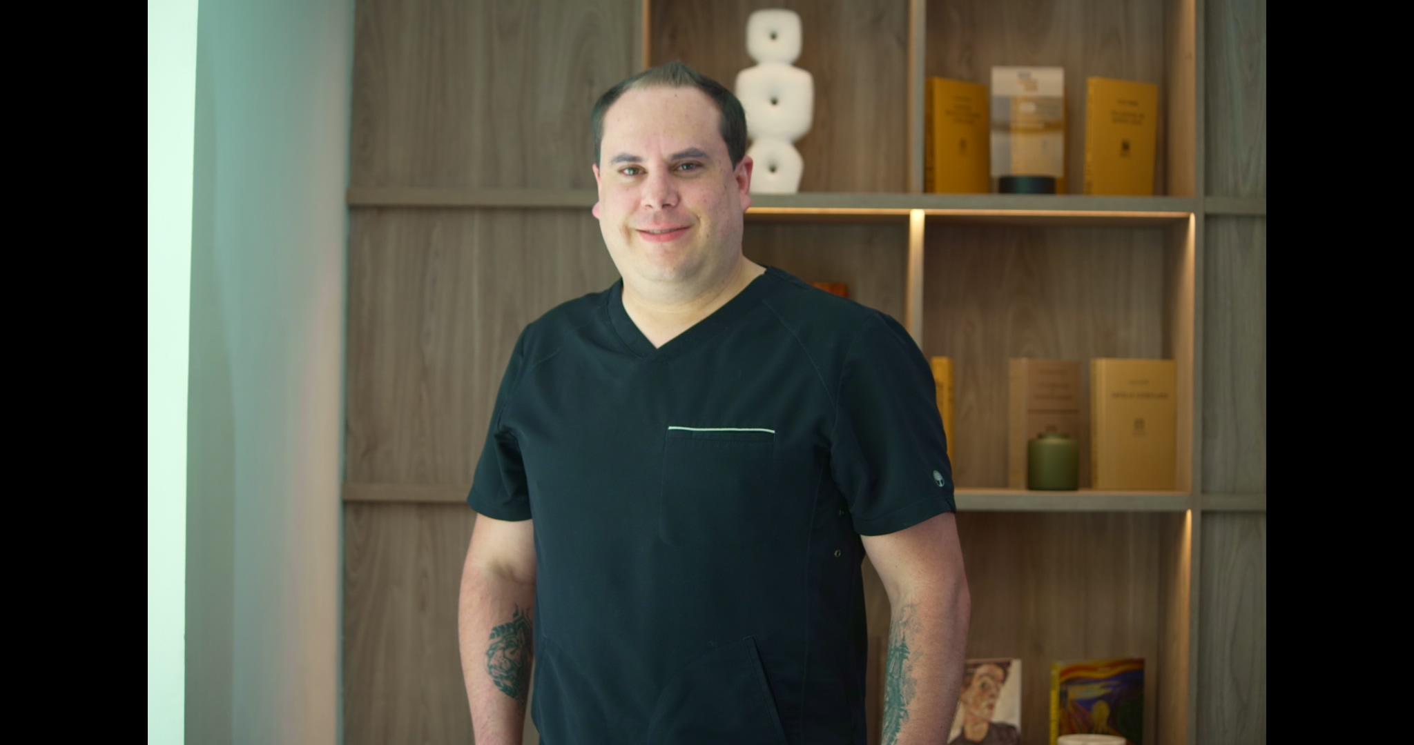 Dr. Javier S in black dental scrubs, smiling confidently in a modern clinic setting with shelves displaying dental materials and books.