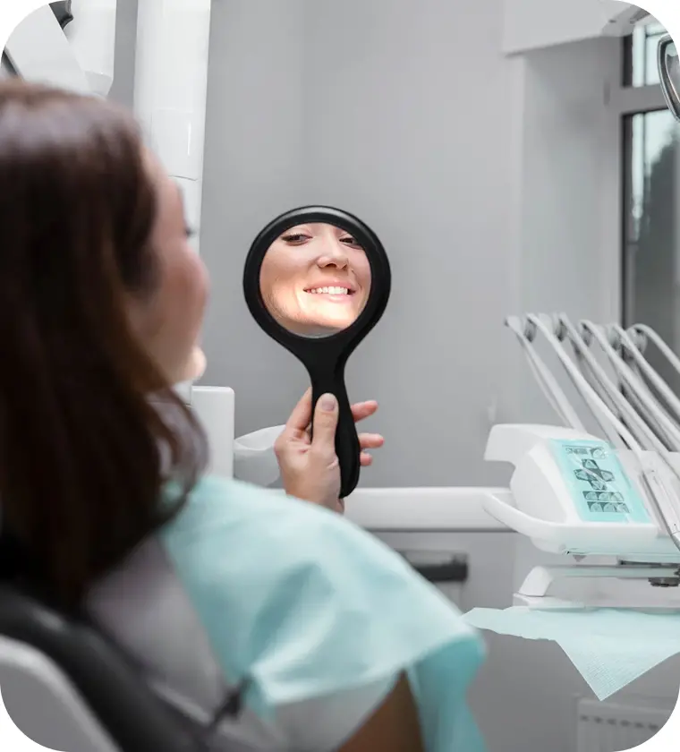 Woman smiling while holding a dental mirror in a clinic, reflecting a positive dental experience, emphasizing patient satisfaction in dental treatments.