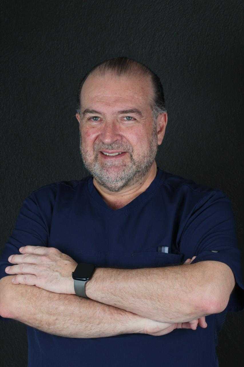 Dr. Sergio M. in dental scrubs, smiling with arms crossed, showcasing expertise in periodontics and implantology, against a dark background.